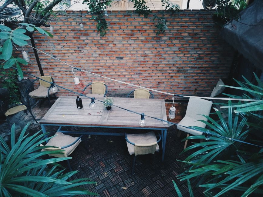 An overhead view of a small patio yard featuring a rectangular wooden dining table with a weathered surface, surrounded by six beige cushioned chairs with dark metal frames. The patio is enclosed by a