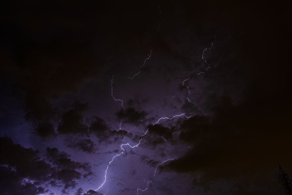 A dark, stormy night sky filled with thick, black clouds illuminated sporadically by sharp, jagged lightning bolts branching across the sky. The lightning reveals faint outlines of clouds and the silh